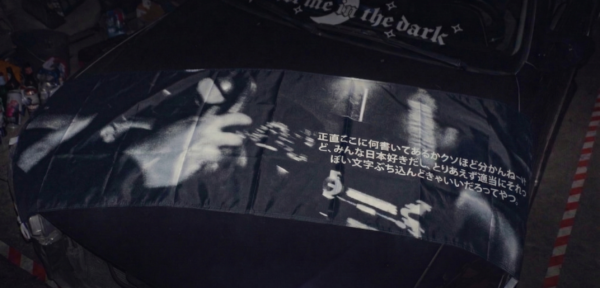 Flaga Nobori "Japońskie pie*dolenie" w stylu JDM, czarno-biała, rozłożona na masce ciemnego samochodu w garażu, z widocznym japońskim tekstem.